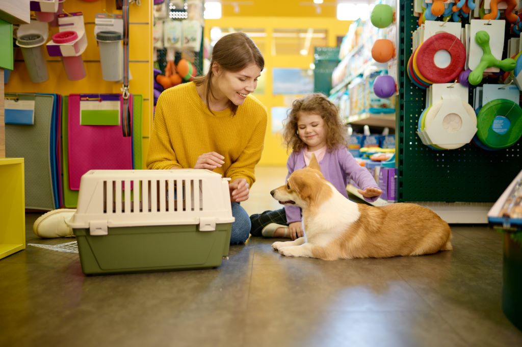 Na foto, há uma criança vestida com um sueter lilás, passando a mão em um cãozinho em sua frente, ao seu lado está uma mulher com sueter mostarda e ambas estão dentro de um pet-shop.